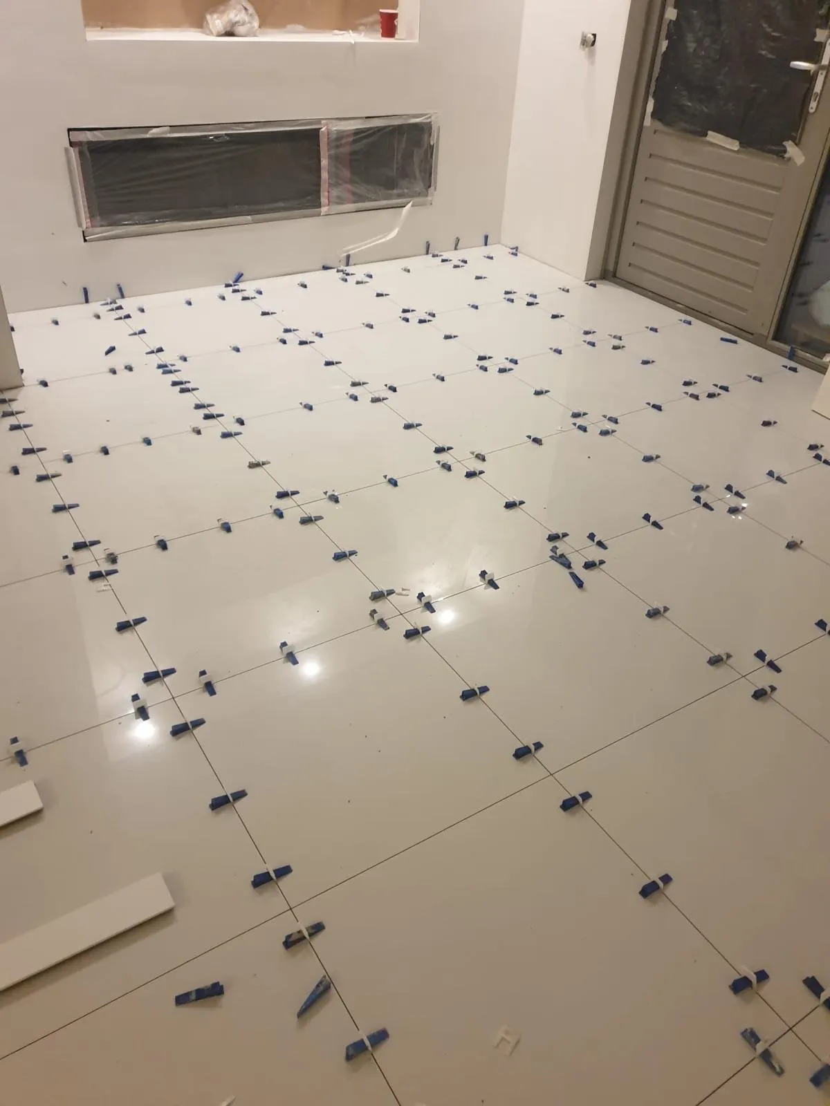 Grote lichte vloertegels tijdens het leggen, met blauwe nivelleerclips tussen de voegen